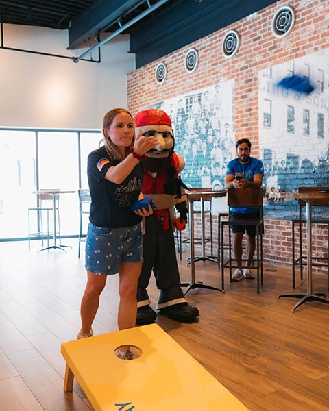 Eine Frau wirft ein blaues Säckchen beim Cornhole-Spiel in einer Bar neben einem Piraten-Maskottchen.