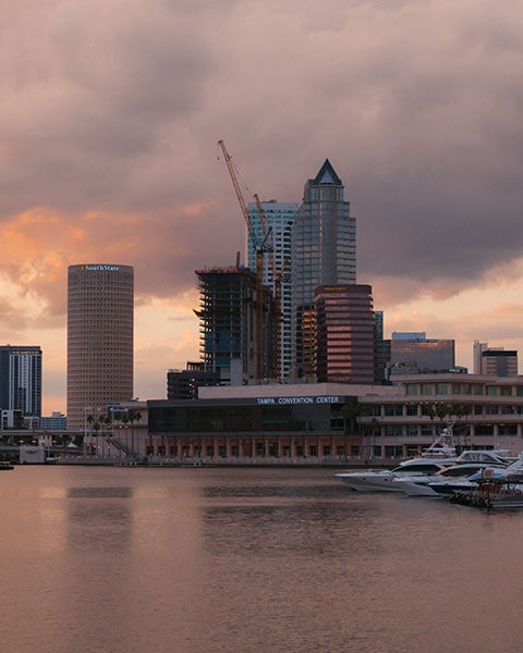 Das Tampa Convention Center und die dahinterliegende Skyline mit Baustellenkranen spiegeln sich bei Sonnenuntergang im Wasser.