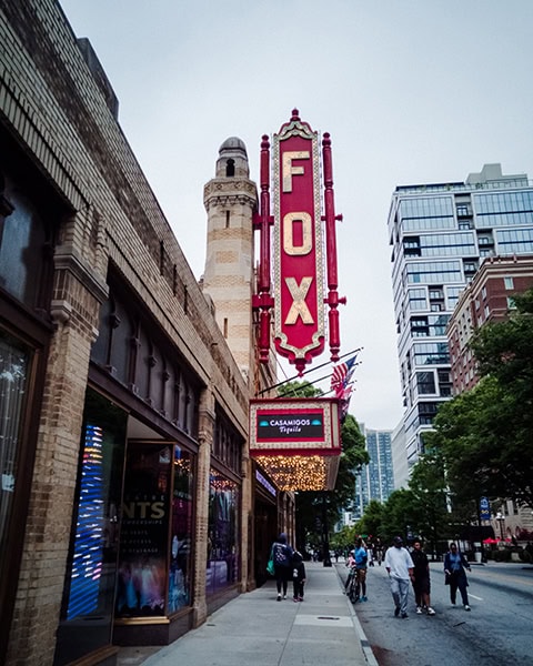Ein großes, rotes, vertikales Neon-Schild mit der Aufschrift „FOX“ ragt an der Fassade eines historischen Theaters im maurischen Baustil empor, während Passanten auf dem Gehweg davor entlanggehen.