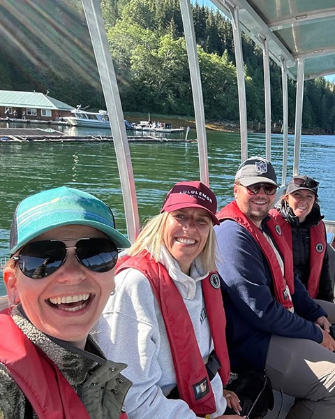 Vier lächelnde Personen (drei Frauen und ein Mann) sitzen nebeneinander auf einem überdachten Boot im Sonnenschein. Sie tragen rote Rettungswesten, Kappen und Sonnenbrillen. Im Hintergrund ist grünes Fjordwasser zu sehen, dahinter ein bewaldetes Ufer mit einer Lodge und angedockten Booten.