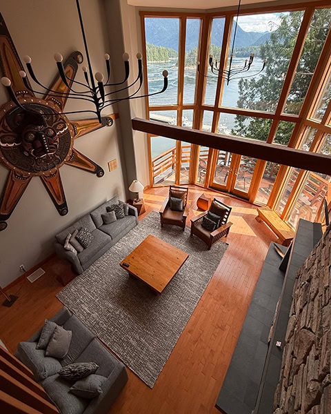Blick von oben in eine riesige, moderne Hotel-Lobby mit hohen Decken und einer riesigen Panorama-Glasfront, die den Blick auf den Ozean freigibt. Im Raum stehen graue Sofas, Ledersessel und ein Holztisch auf einem großen Teppich. An der Wand links hängt eine monumentale, kunstvoll geschnitzte Sonnenmaske im traditionellen Stil der First Nations.