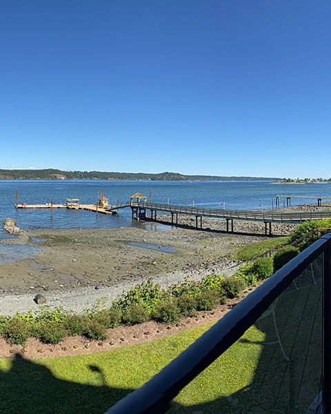 Blick von einem Balkon auf einen langen Holzsteg, der über einen kiesigen Strand zu einer schwimmenden Bootsanlegestelle im tiefblauen Meer führt. Im Hintergrund erstreckt sich eine bewaldete Landzunge unter wolkenlosem blauem Himmel, während im Vordergrund gepflegter grüner Rasen zu sehen ist.