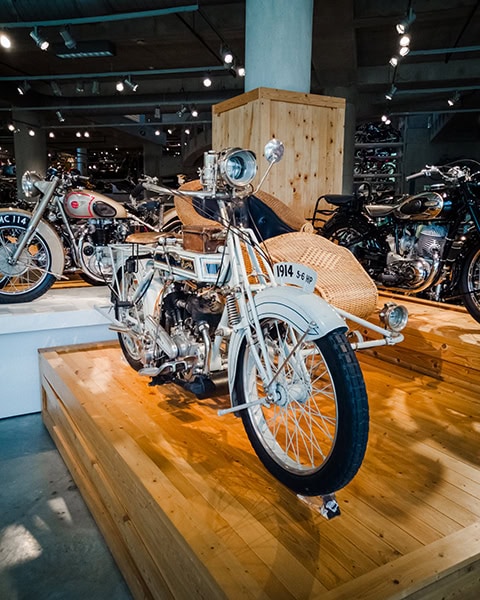 Ein historisches, weißes Motorrad aus dem Jahr 1914 mit einem geflochtenen Beiwagen aus Korbweide steht auf einem Holzpodest inmitten einer Ausstellung mit weiteren Oldtimer-Motorrädern unter Deckenstrahlern.