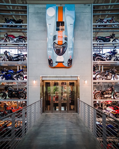 Ein hellblauer Porsche 917 Rennwagen im legendären orange-blauen Gulf-Design hängt vertikal über einer Glastür an einer Betonwand, flankiert von mehrstöckigen Regalen voller historischer Motorräder in einem großen Museum.