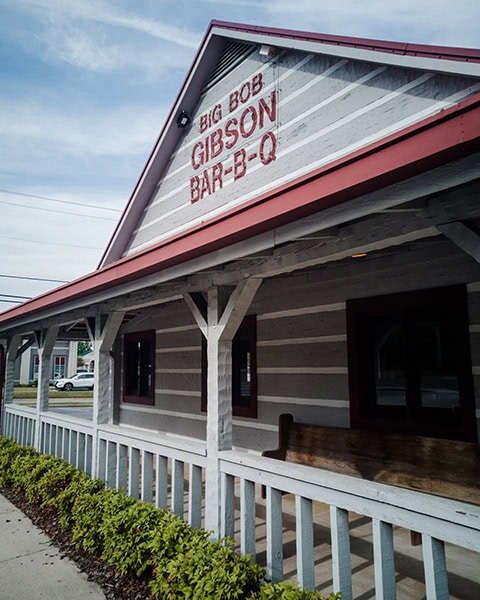 Die Außenansicht eines rustikalen Holzgebäudes mit einer überdachten Veranda zeigt im oberen Giebel den roten Schriftzug „BIG BOB GIBSON BAR-B-Q“ auf weiß gestrichenem Holz unter einem leicht bewölkten Himmel.