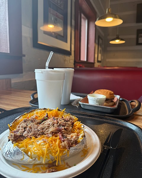 Eine große, in Alufolie gewickelte Ofenkartoffel ist reichlich mit geriebenem Käse und Pulled Pork belegt und steht auf einem Tablett im Vordergrund, während im Hintergrund ein Burger und zwei weiße Becher in einem Restaurantbereich zu sehen sind.