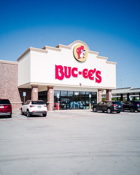 Die große, helle Außenfassade einer Buc-ee's-Filiale mit dem markanten roten Schriftzug und dem runden Biber-Logo steht hinter einem Parkplatz mit mehreren Autos unter einem strahlend blauen Himmel.