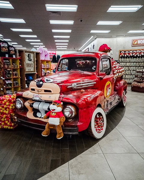 Ein Oldtimer-Lieferwagen in rotem Vintage-Look steht als Dekorations- und Verkaufsfläche inmitten eines hell erleuchteten Buc-ee’s Reisemarktes; er ist mit dem Biber-Maskottchen bemalt und mit zahlreichen Plüschtieren beladen.
