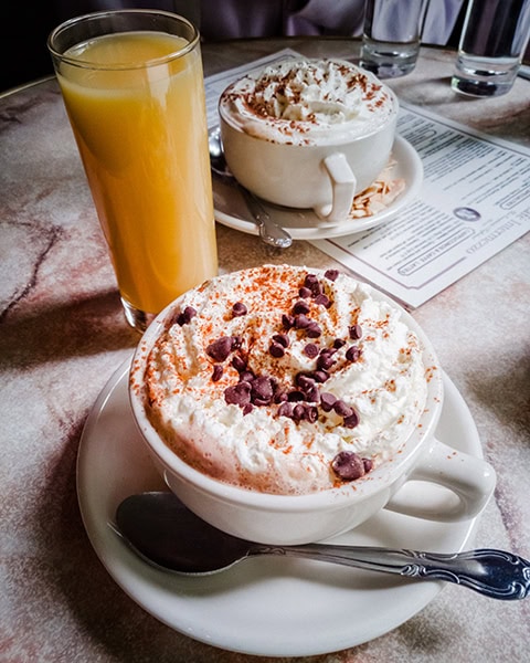 Ein großes Glas Orangensaft und zwei Kaffeetassen mit reichlich Schlagsahne, Kakaopulver und Schokoladenstückchen stehen zusammen mit Löffeln auf einem Tisch im Café Intermezzo in Atlanta.