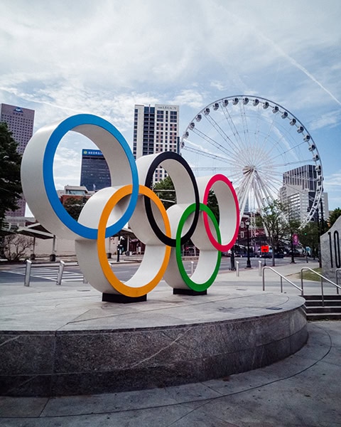 Eine große, dreidimensionale Skulptur der fünf farbigen Olympischen Ringe steht auf einem Steinsockel im Centennial Olympic Park, während im Hintergrund ein großes, weißes Riesenrad und Wolkenkratzer unter einem leicht bewölkten Himmel zu sehen sind.