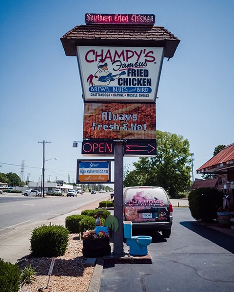 Ein großes, mehrteiliges Werbeschild für „Champy's Famous Fried Chicken“ steht an einer Straße, zeigt ein Huhn mit Sonnenbrille und Gitarre sowie eine LED-Anzeige mit der Aufschrift „Always fresh & hot“ über einem geparkten Auto und einer blauen Toilette als Dekoration am Straßenrand.
