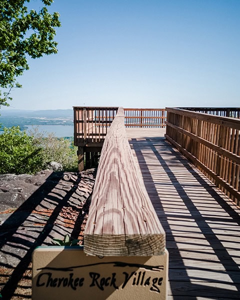 Eine hölzerne Aussichtsplattform mit Geländer führt zu einem Aussichtspunkt auf einer Anhöhe, von dem aus man im Hintergrund einen weiten Blick auf einen See und Hügel unter blauem Himmel hat; im Vordergrund ist ein Schild mit der Aufschrift „Cherokee Rock Village“ zu sehen.