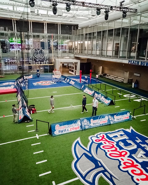 Ein Mini-Footballfeld im Innenbereich der College Football Hall of Fame in Atlanta ist mit einem künstlichen Rasen, Linienmarkierungen und Werbebanden für die „Chick-fil-A Peach Bowl Skillzone“ ausgestattet, während einige Personen auf dem Feld stehen.