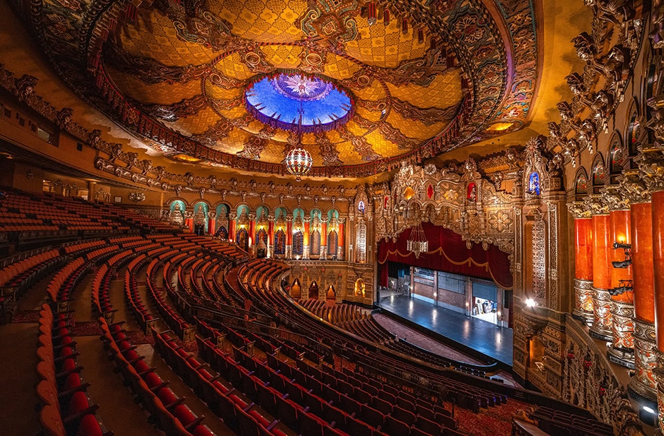 Blick von den oberen Zuschauerrängen in den prachtvollen, riesigen Saal des historischen Fox Theatre in Detroit. Die Architektur ist im opulenten, kambodschanisch-indischen Stil mit reich verzierten, goldenen Säulen, roten Akzenten und Skulpturen gestaltet. Eine gewaltige, goldene Kuppeldecke mit einem zentralen, leuchtend blauen Kreis und einem prunkvollen Kronleuchter überspannt die roten Kinosessel, die sich im Halbkreis um die beleuchtete Bühne mit rotem Vorhang anordnen.