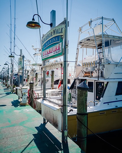 Ein grüner Steg führt an einer Reihe von angedockten Fischerbooten und Yachten in der HarborWalk Marina in Destin, Florida, vorbei, wobei im Vordergrund ein großes Schild für das Charterboot „STARFIRE“ unter historischen Straßenlaternen und einem klaren blauen Himmel zu sehen ist.