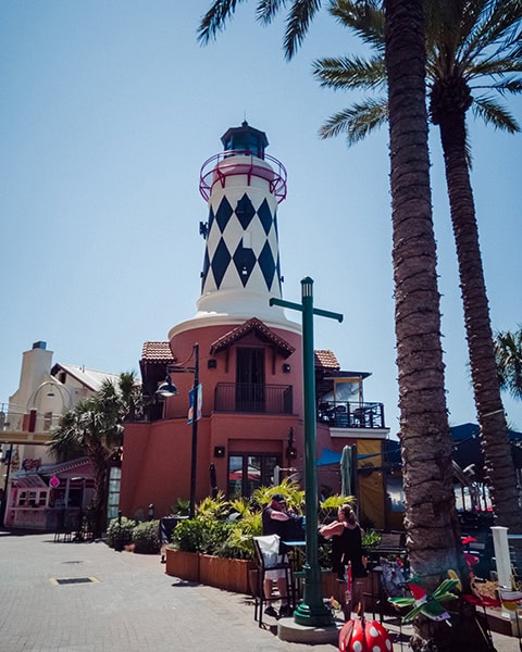 Ein markanter Leuchtturm mit einem schwarz-weißen Rautenmuster ragt über dem terracottafarbenen Gebäude des Restaurants Margaritaville im HarborWalk Village in Destin, Florida, auf, während im Vordergrund Palmen und ein gepflasterter Fußgängerbereich mit Außengastronomie unter klarem blauem Himmel zu sehen sind.