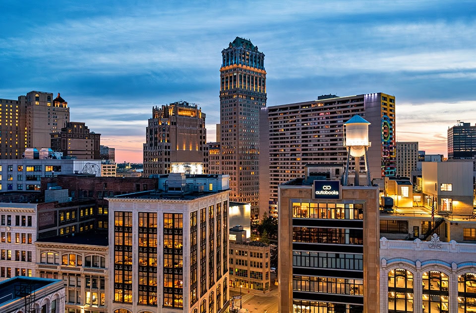Eine erhöhte Stadtansicht von Downtown Detroit während der Dämmerung zur blauen Stunde. Im Vordergrund sind beleuchtete historische und moderne Gebäude zu sehen, darunter eines mit einem beleuchteten Wasserturm und dem Logo ‚autobooks‘ auf dem Dach. Im Hintergrund ragt der markante, kunstvoll illuminierte Book Tower empor, umgeben von weiteren Hochhäusern unter einem blau-orange gefärbten Abendhimmel.