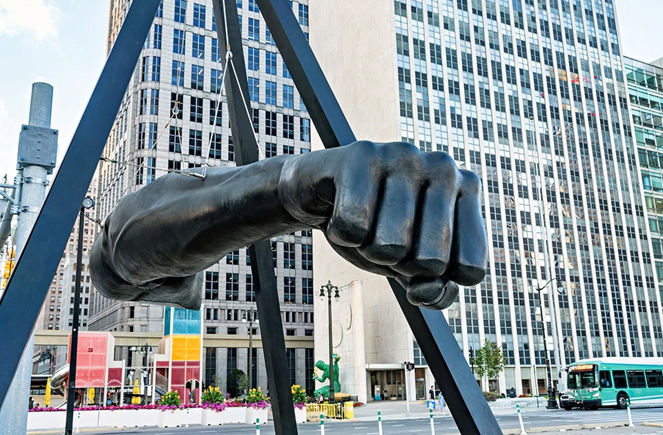 Eine riesige, schwarze Bronze-Skulptur einer geballten Faust, die an einem pyramidenartigen Metallgerüst aufgehängt ist. Die Skulptur, bekannt als Monument für Joe Louis, befindet sich im Zentrum von Detroit. Im Hintergrund ragen moderne Bürogebäude und Hochhäuser empor, rechts ist ein grüner Stadtbus zu sehen.