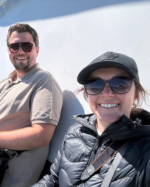 Ein lächelnder Mann und eine lächelnde Frau sitzen nebeneinander auf dem Außendeck einer Fähre im Sonnenschein. Beide tragen Sonnenbrillen; die Frau im Vordergrund trägt eine schwarze Daunenjacke und eine schwarze Basecap mit einem gestickten Ahornblatt.