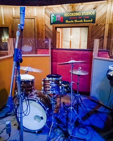 Ein aufgebautes, holzgemustertes Schlagzeug mit Becken steht im blau ausgeleuchteten Aufnahmebereich eines Musikstudios, während im holzgetäfelten Hintergrund ein Schild mit der Aufschrift „FAME RECORDING STUDIOS - HOME OF THE Muscle Shoals Sound“ über einem Studiofenster hängt.