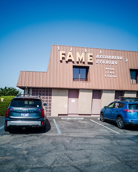 Die Außenansicht eines zweistöckigen Gebäudes mit brauner Fassade zeigt den großen Schriftzug „FAME RECORDING STUDIOS“ und den Zusatz „WHERE IT ALL STARTED“, während davor zwei SUVs auf einem asphaltierten Parkplatz unter blauem Himmel stehen.