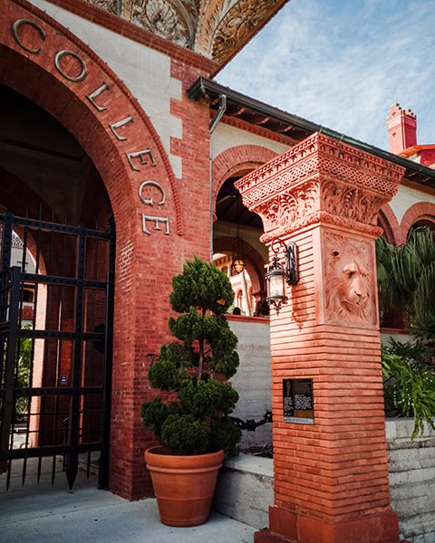 Ein imposanter roter Backsteinbogen mit der eingeprägten Aufschrift „COLLEGE“ markiert den Eingangsbereich zum historischen Hauptgebäude des Flagler College (dem ehemaligen Hotel Ponce de Leon) in St. Augustine, Florida, flankiert von einem verzierten Terrakotta-Pfeiler mit einem eingearbeiteten Löwenkopf, einer historischen Laterne und einem formgeschnittenen Nadelbaum im Tontopf.