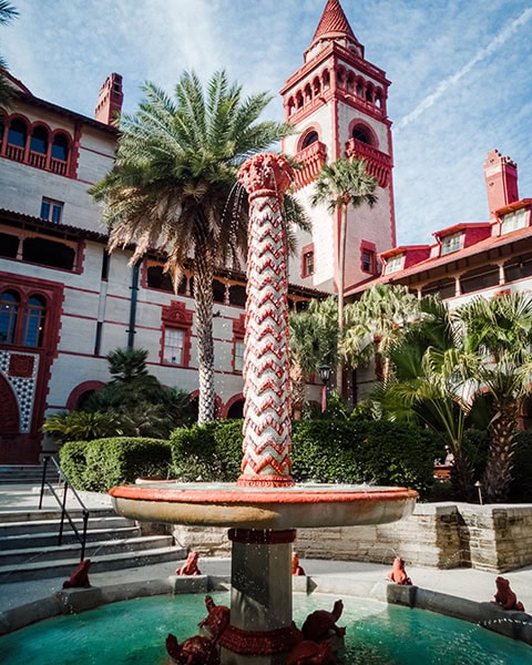 Der kunstvolle Frosch-Springbrunnen (Frog Fountain) plätschert mit seinem rot-weiß gemusterten Mittelpfeiler inmitten des von Palmen und tropischen Pflanzen gesäumten Innenhofs des Flagler College (dem ehemaligen Hotel Ponce de Leon) in St. Augustine, Florida, während im Hintergrund die markanten roten Fensterbögen und einer der großen Glockentürme der historischen Anlage in den leicht bewölkten Himmel ragen.