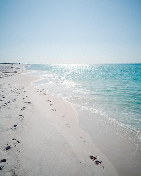 Der weiße, feinkörnige Sandstrand von Rosemary Beach (oder dem direkt angrenzenden Inlet Beach) erstreckt sich entlang des klaren, türkisfarbenen Wassers des Golfs von Mexiko, in dem sich das helle Sonnenlicht spiegelt, während im feuchten Ufersand zahlreiche Fußabdrücke unter einem wolkenlosen, blauen Himmel zu sehen sind.