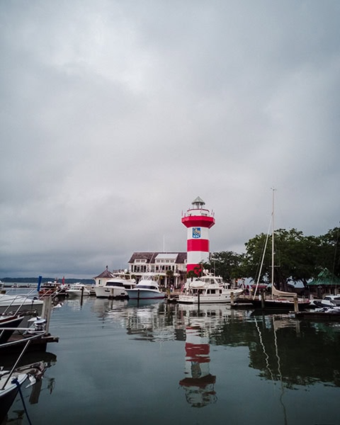 Der rot-weiß gestreifte Harbour Town Lighthouse ragt im Zentrum der Harbour Town Marina auf Hilton Head Island, South Carolina, empor und spiegelt sich zusammen mit mehreren Yachten und Segelbooten auf der ruhigen Wasseroberfläche des Hafens unter einem wolkenverhangenen Himmel.