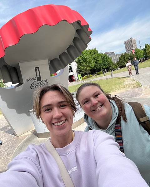 Zwei lächelnde junge Frauen machen ein Selfie im Freien vor einer großen Skulptur in Form eines roten Coca-Cola-Flaschendeckels mit dem Schriftzug „World of Coca-Cola“.