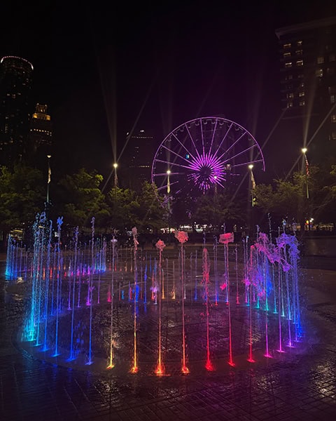 Ein beleuchteter Springbrunnen mit bunten, kreisförmig angeordneten Wasserfontänen sprüht bei Nacht im Centennial Olympic Park, während im dunklen Hintergrund ein lila erleuchtetes Riesenrad und die Umrisse von Wolkenkratzern zu sehen sind.