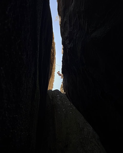 Der Blick aus einer tiefen, dunklen Felsspalte nach oben zeigt einen Kletterer, der an Seilen gesichert an einer engen, hell erleuchteten Felsöffnung emporsteigt.