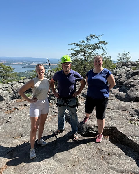 Drei lächelnde Personen stehen nebeneinander auf einem felsigen Berggipfel, wobei der Mann in der Mitte einen hellgrünen Kletterhelm und Klettergurt trägt, während sich im Hintergrund eine weite Landschaft mit einem großen See erstreckt.