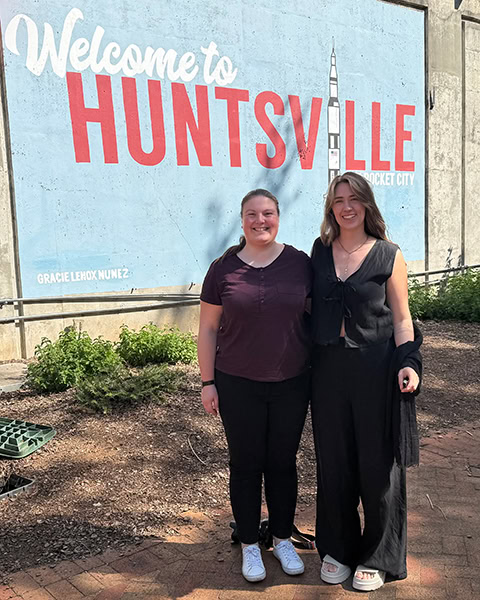 Zwei lächelnde junge Frauen stehen nebeneinander vor einer großen Wandmalerei mit der Aufschrift „Welcome to HUNTSVILLE - Rocket City“, auf der auch eine stilisierte Rakete abgebildet ist.