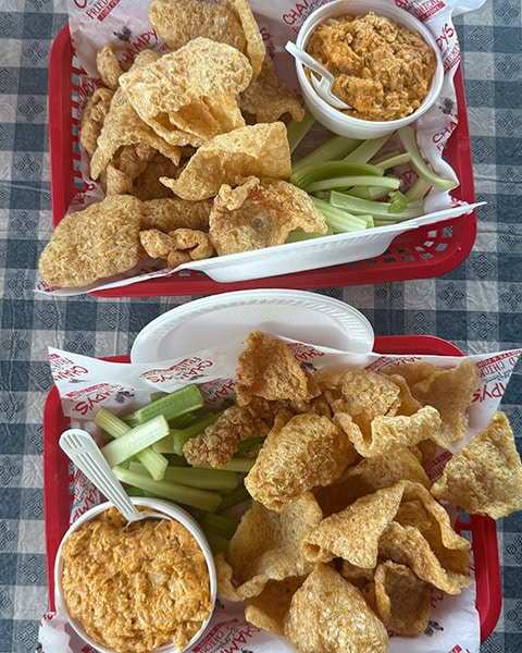 Zwei rote Servierkörbe stehen auf einer blau-weiß karierten Tischdecke, jeweils gefüllt mit einer großen Portion knuspriger Schweineschwarten (Pork Rinds), frischen Selleriestangen und einem Becher cremigen, orangefarbenen Buffalo-Chicken-Dips mit Plastiklöffeln.