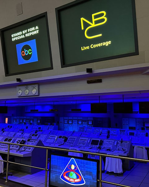 Ein historischer Kontrollraum im Kennedy Space Center mit blau beleuchteten Konsolen, Bildschirmen von ABC und NBC sowie einem Apollo-8-Emblem.