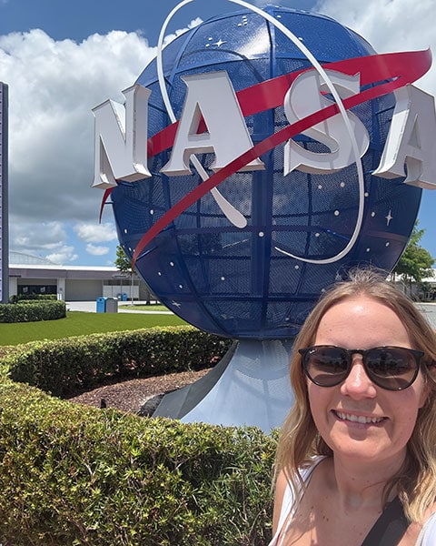Eine lächelnde Frau mit Sonnenbrille macht ein Selfie vor dem großen, blauen NASA-Logo am Kennedy Space Center.