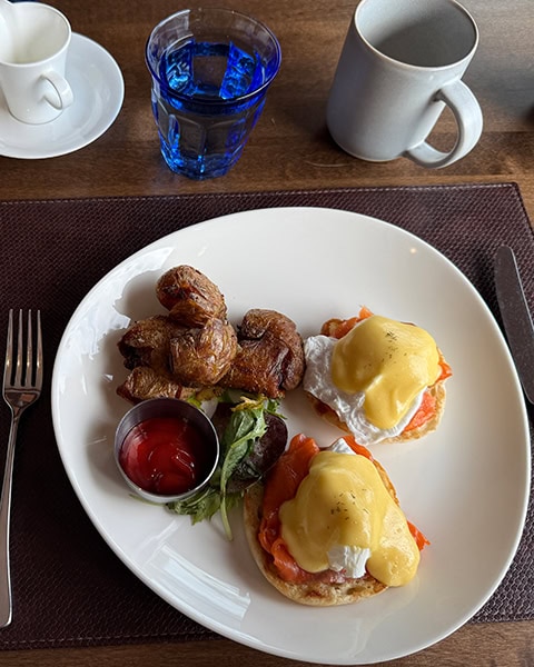 Ein weißer Teller auf einem braunen Platzset zeigt zwei pochierte Eier Benedict mit Räucherlachs auf englischen Muffins, überzogen mit Sauce Hollandaise. Daneben liegen goldbraun geröstete Kartoffeln, eine kleine Schale Ketchup und etwas Salat. Im Hintergrund stehen ein blaues Wasserglas sowie Kaffeetassen auf dem Holztisch.