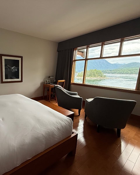 Ein helles Hotelzimmer mit glänzendem Holzboden und einem großen Bett mit weißer Bettwäsche im Vordergrund. Zwei dunkelgraue Sessel sind zu einem großen Holzfenster hin ausgerichtet, das einen freien Blick auf ein ruhiges Gewässer, eine kleine bewaldete Insel und majestätische Berge im Hintergrund bietet.