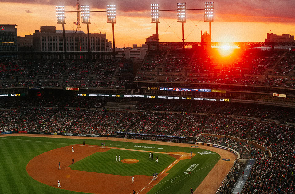 Blick von einer erhöhten Tribüne auf ein gut besuchtes Baseballstadion, den Comerica Park in Detroit, während eines Spiels. Die tiefstehende Sonne bricht dramatisch durch die oberen Zuschauerränge im Hintergrund und taucht den Himmel in ein intensives Orange-Rot. Große Flutlichtmasten ragen in den Abendhimmel, während unten auf dem grünen Rasen und der Sandfläche das Spiel läuft.