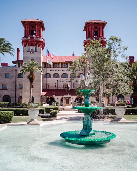 Ein grüner, mehrstufiger Springbrunnen plätschert im großen Wasserbecken direkt vor der historischen Palastfassade des Lightner Museum (dem ehemaligen Hotel Alcazar) in St. Augustine, Florida, flankiert von zwei markanten, rötlichen Türmen, Palmen und einer bronzenen Statue im gepflegten Museumsgarten unter klarem blauem Himmel.