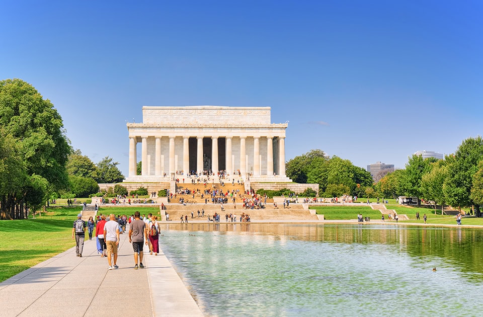 Frontalansicht des weißen, klassizistischen Lincoln Memorials unter einem klaren, strahlend blauen Sommerhimmel. Auf den monumentalen Treppenstufen befinden sich viele Touristen. Im Vordergrund erstreckt sich der Lincoln Memorial Reflecting Pool mit hellem, leicht grünlichem Wasser. Auf dem gepflasterten Gehweg links spazieren mehrere Menschen in Sommerkleidung in Richtung des Denkmals. Die umliegenden Bäume und Rasenflächen sind saftig grün.