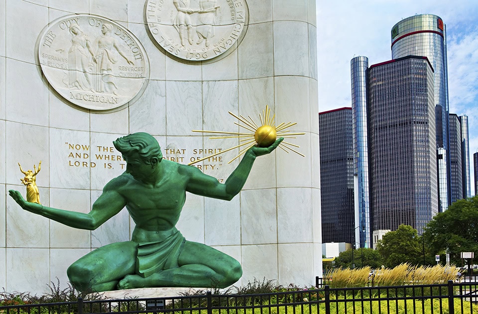 Die ikonische grüne Bronzestatue ‚The Spirit of Detroit‘ sitzt vor einer weißen Marmorwand mit den Siegeln der Stadt Detroit und des Bundesstaates Michigan. In der rechten Hand hält die Statue eine kleine goldene Familiengruppe, in der linken Hand eine goldene Kugel mit Strahlen. Im Hintergrund ragen die modernen, verglasten Wolkenkratzer des Renaissance Center empor.