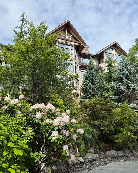 Blick nach oben auf ein mehrstöckiges, rustikales Hotelgebäude mit großen Glasfronten und Giebeln aus Holz. Das Gebäude ist von dichten Nadelbäumen umgeben. Im Vordergrund blühen hellrosa Rhododendronbüsche an einem steinigen Hang unter einem leicht bewölkten Himmel.