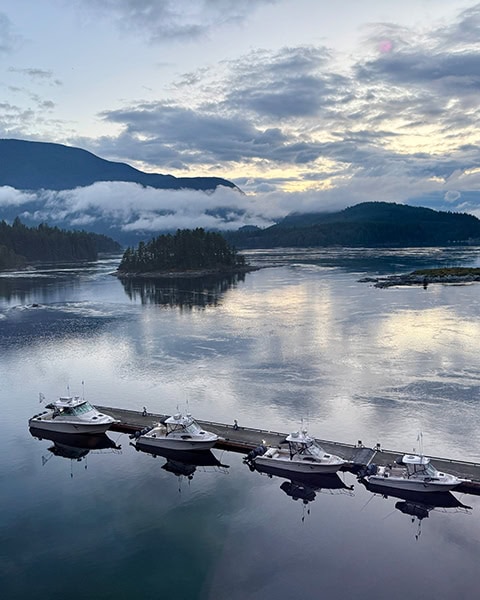 Alt-Text: Blick von oben auf eine ruhige Bucht bei Dämmerung. An einem langen Holzsteg liegen vier weiße Motorboote im spiegelglatten Wasser. Mittig im Wasser liegt eine kleine, bewaldete Insel. Im Hintergrund erstrecken sich bewaldete Berge, um die tiefe Nebelschwaden hängen, unter einem bewölkten Abendhimmel.
