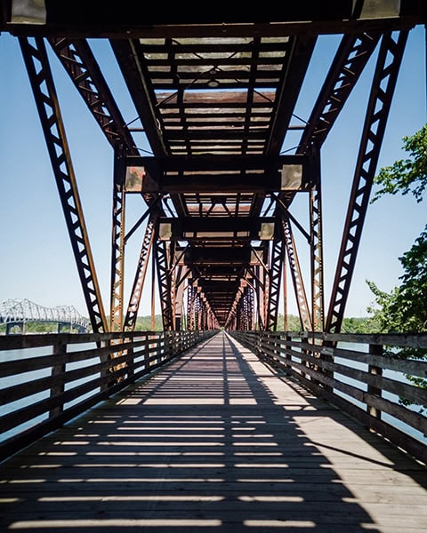 Der Blick entlang einer langen Fußgängerbrücke aus Holzbohlen führt durch eine massive, rostige Stahlträgerkonstruktion, die im Sonnenlicht lange Schatten wirft, während links eine weitere Brücke über dem Fluss zu sehen ist.