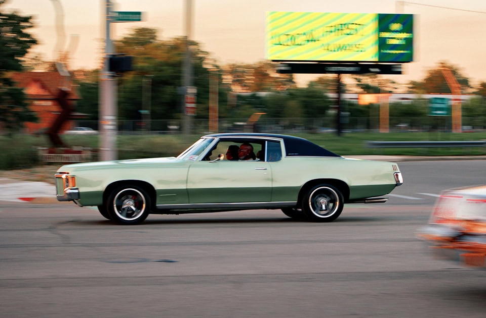 Ein klassisches, hellgrünes Pontiac Grand Prix Coupé aus den frühen 1970er-Jahren mit schwarzem Vinyldach fährt im Abendlicht über eine Straße in Detroit. Im unscharfen Hintergrund sind Bäume, eine Ampel und eine große leuchtende Plakatwand zu sehen. Die Bewegung des Autos erzeugt eine leichte Bewegungsunschärfe