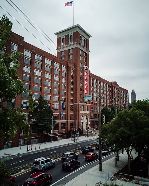 Ein riesiger, historischer Backsteinkomplex mit einem markanten Turm und einem vertikalen Leuchtschild mit der Aufschrift „PONCE“ steht an einer belebten Straße mit fließendem Autoverkehr unter einem bewölkten Himmel.
