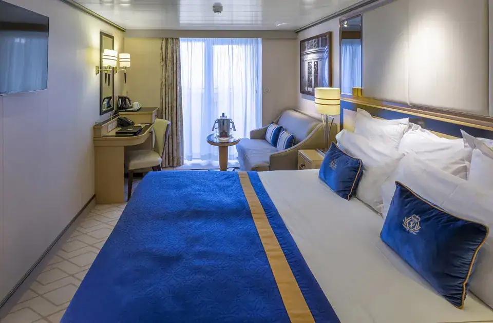 Das Innere einer eleganten Kreuzfahrtkabine mit einem großen Doppelbett, das mit einer königsblauen Tagesdecke und passenden Kissen mit goldenem Logo dekoriert ist. Im Hintergrund befinden sich ein kleines Sofa, ein kleiner Tisch mit einem Sektkühler und eine Balkontür mit hellen Vorhängen. Links an der Wand sind ein Schreibtisch mit einem Stuhl und ein Fernseher angebracht.