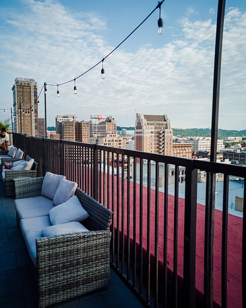 Eine Reihe von grauen Lounge-Sofas aus Rattan mit hellen Kissen steht entlang des Geländers einer Dachterrasse, über der eine Lichterkette gespannt ist, während sich im Hintergrund die Skyline einer Stadt mit historischen Hochhäusern unter leicht bewölktem Himmel erstreckt.